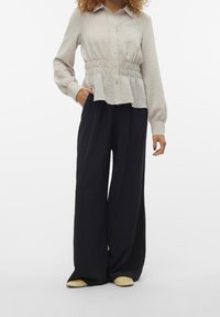 Camicia grigia chiaro, con texture e bottoni, con vita elastica; abbinata a pantaloni neri a gamba larga e scarpe gialle chiare senza lacci.
