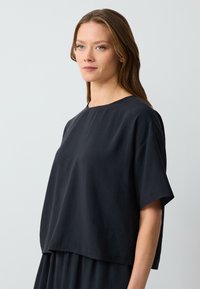 Top nero a maniche corte, realizzato in tessuto morbido, con scollatura rotonda e una vestibilità ampia e svasata, che arriva fino ai fianchi.