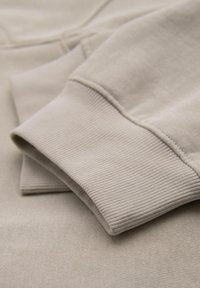Béžová bavlněná látka na mikinu s hladkým povrchem a žebrovanými manžetami, prezentující složený design a jemnou strukturu.