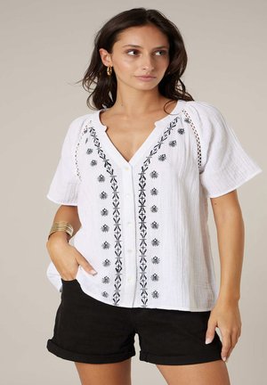 Femme portant un chemisier blanc à manches courtes avec broderie noire et des shorts noirs à ourlet roulé, posant avec une main dans la poche.