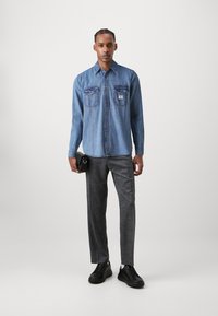 Denimhemd, hellblau, mit Knöpfen und zwei Brusttaschen; kombiniert mit grauen Hosen und schwarzen Sneakers, gehalten mit einer kleinen schwarzen Tasche.