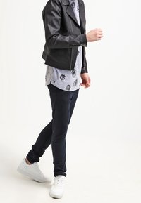 Black leather biker jacket over a gray floral-patterned shirt, paired with dark jeans and white sneakers. Actor is in a walking pose.