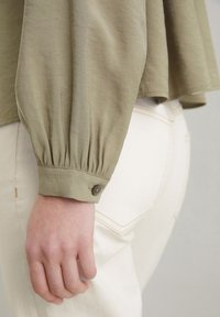 Olijfgroen overhemd met lange mouwen en elastische manchetten, gecombineerd met lichtgekleurde broek met een achterzak en zichtbare stiksels.