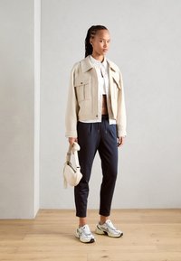 Beige cropped jack met grote zakken, wit overhemd, donker taps toelopende broek en zilveren sneakers; draagt een kleine crèmekleurige handtas.