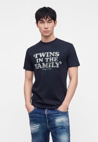Νέος άνδρας με μαύρα μαλλιά φοράει μαύρο T-shirt με κείμενο «"Twins in the Family"» και φθαρμένο μπλε τζιν, χέρια στις τσέπες, ουδέτερο φόντο.