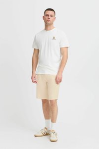 Fehér pamut póló logóval, beige rövidnadrág, fehér zokni és világos sportcipő. A modell lazán áll, karjai pihentetve, semleges háttérrel.