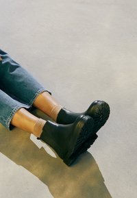 Schwarze Leder-Ankle-Boots mit strukturierten Sohlen, kombiniert mit beigen gerippten Socken, auf einer hellen Betonoberfläche; Jeanshosen teilweise sichtbar.