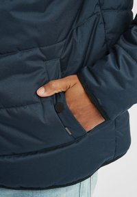 Navy puffer jacket featuring a zippered pocket, textured fabric, and a round black button. The hem is adjustable with a black trim.