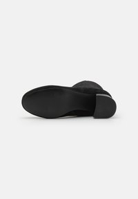Botte à talon en daim noir avec un bout arrondi, un dessus texturé et une semelle plate en caoutchouc. Le talon est court et de forme bloc.
