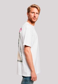 Weißes Baumwoll-T-Shirt mit kurzen Ärmeln, das ein buntes Grafikdesign auf dem Rücken zeigt, und einem lässigen Schnitt. Dazu werden Denim-Jeans getragen.