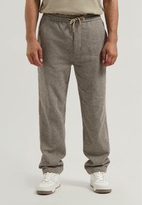 Man gekleed in losse broek met een bruin en grijs houndstooth patroon, met een trekkoord in de taille en witte sneakers, staande tegen een effen achtergrond.