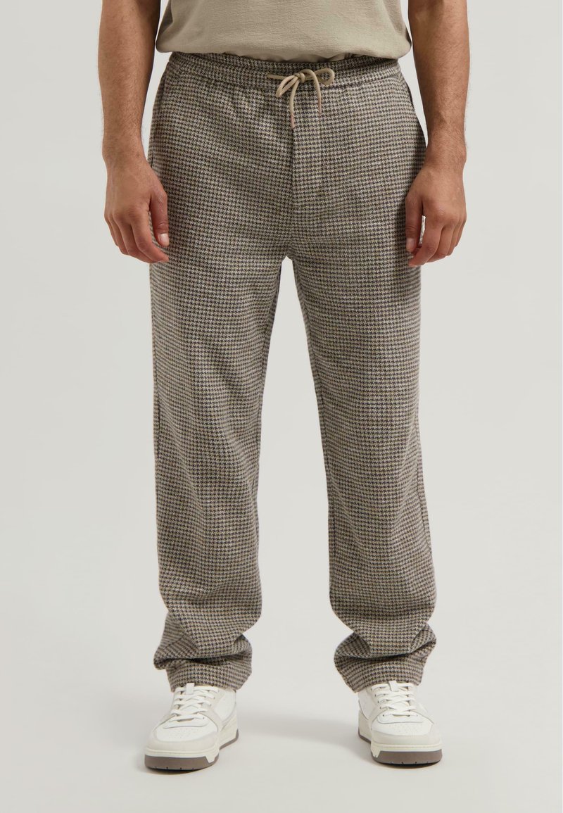Man gekleed in losse broek met een bruin en grijs houndstooth patroon, met een trekkoord in de taille en witte sneakers, staande tegen een effen achtergrond.