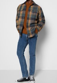 Rutig flanellskjorta i blått, brunt och grönt, med knäppta bröstfickor, lager över en brun tröja och blå jeans. Svarta sneakers.