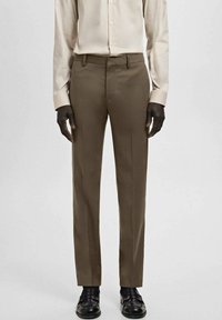 Braune, maßgeschneiderte Hose aus glattem Stoff mit schmaler Passform, Gürtelschlaufen und gebügelten Biesen an der Vorderseite. Kombiniert mit einem hellen Hemd.