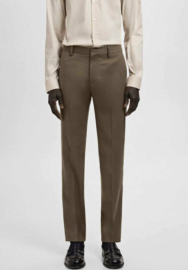Braune, maßgeschneiderte Hose aus glattem Stoff mit schmaler Passform, Gürtelschlaufen und gebügelten Biesen an der Vorderseite. Kombiniert mit einem hellen Hemd.