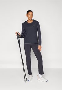 Zwarte lange mouwtop met een ronde halslijn, ritszakken, gecombineerd met donkere broek. Het model houdt een trekkingstok vast en draagt lichte sportschoenen.