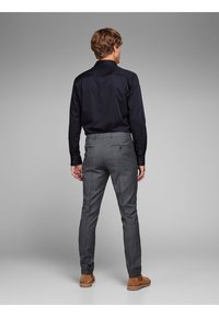 Man in een donkerblauwe overhemd met lange mouwen en grijs geblokte broek, gezien vanaf de achterkant. Bruine schoenen en subtiele merkdetails op de broek.