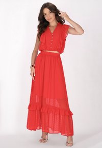 Top corto rosso a balze con bottoni e gonna lunga coordinata con balze sovrapposte e sezioni trasparenti; tessuto leggero e testurizzato.