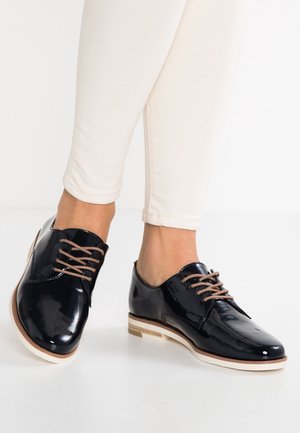 Sorte laklæder snørebåndssko med rund tå, beige snørebånd og en kontrastfyldt hvid sål med brune accenter.