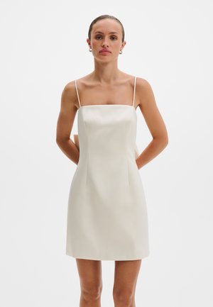 Žena oblečená v bezrukávových, po kolená siahajúcich slonovinových saténových šatách s tenkými ramienkami, stojaca pred jednoduchým bielym pozadím.