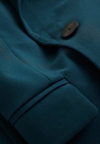 Blågrön tyg med en slät textur, utrustad med en framträdande svart knapp och detaljerad sömnad längs kanterna. Närbild av ett plagg.