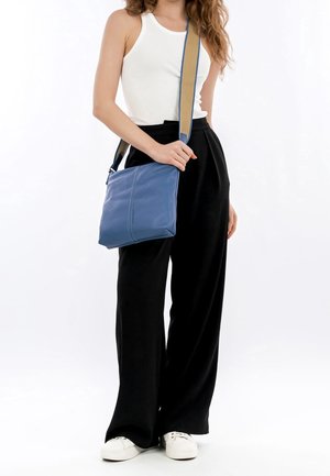 Donna che indossa un top bianco a canotta, pantaloni neri a gamba larga, sneakers bianche, con una borsa a tracolla blu con tracolla beige su sfondo bianco.