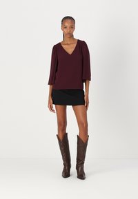 Burgundijska bluza s V-izrezom i napuhanih rukavima, u kombinaciji s crnom mini suknjom i smeškim kožnim kaubojskim čizmama s složenim vezom.