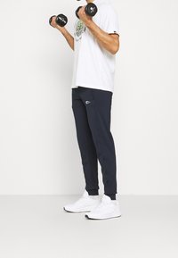 Mann in weißen Adidas-Sneakern, marineblauen Jogginghosen und einem weißen T-Shirt, der 3 kg schwere schwarze Hanteln vor einem weißen Hintergrund hebt.