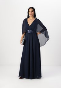 Navy blue long gown with flowing cape sleeves, beaded waistband, V-neck design, and slit detail. Model holds a silver clutch.