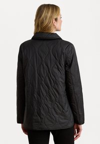 Veste matelassée noire avec un col, présentant un motif de texture ondulée et des manches longues. La vue de dos montre une coupe droite et aucune ornementation visible.