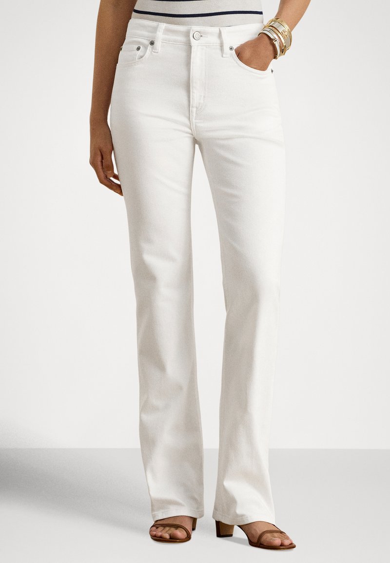 Γυναίκα που φοράει λευκό τζιν με ψηλή μέση και ίσια γραμμή, καφέ πέδιλα με ανοιχτά δάχτυλα και χρυσά βραχιόλια, στέκεται με το ένα χέρι στην τσέπη.