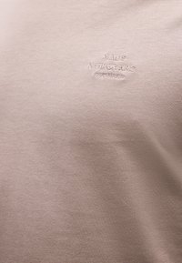 Ανοιχτό μπεζ ύφασμα με μικρό κεντημένο κείμενο που γράφει "MAOS NØRGAARD COPENHAGEN" με κλωστή στο ίδιο χρώμα.