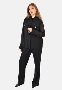 Blusa nera con due tasche sul petto, chiusura con bottoni e maniche lunghe trasparenti, abbinata a pantaloni neri su misura e tacchi neri a punta.