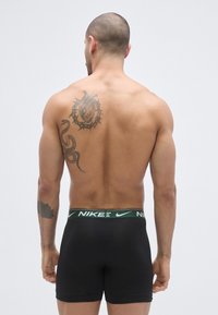 Boxeri negri cu talie elastică, având logo-ul Nike verde. Modelul are un tatuaj pe spate, reprezentând o inimă înconjurată de țepi și un șarpe.