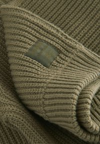 Stickad tröja i olivgrön med texturerad ribbad struktur, vikt för att visa ett tygmärke. Materialet verkar mjukt och något texturerat.
