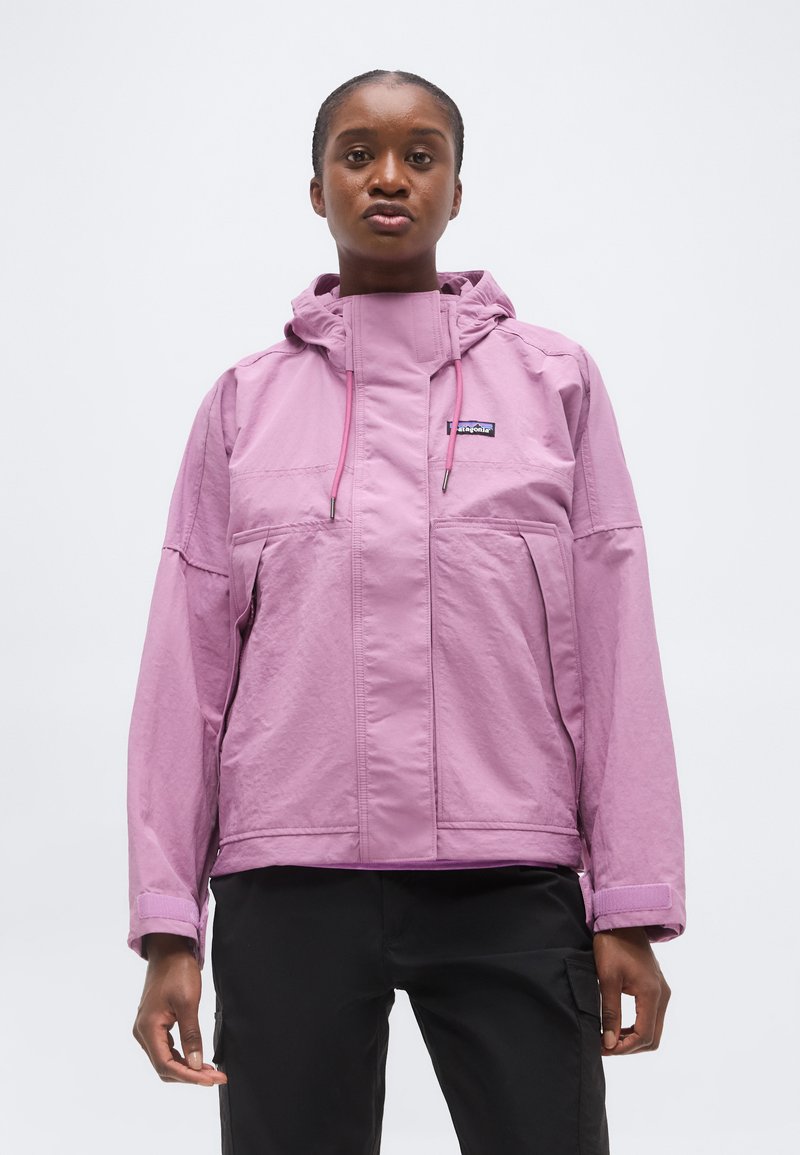 Femme portant une veste à capuche violet clair avec des poches avant et un pantalon noir, debout devant un fond blanc uni.