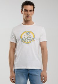 Balta medvilninė marškinė su mėlynu ir geltonu grafiniu dizainu bei užrašu "BIG STAR", apvaliu kaklu ir trumpomis rankovėmis, derinami su mėlynomis džinsomis.