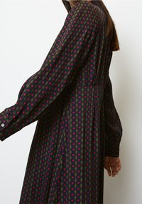 Robe à manches longues en tissu sombre avec un motif géométrique de diamants rouges et verts. Présente une coupe décontractée et des poignets boutonnés.