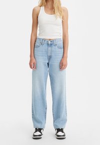 Kvinna klädd i en vit ärmlös crop top, ljusblå högmidjade vida jeans och svartvita sneakers, stående mot en vit bakgrund.