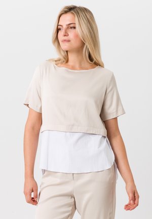 Haut beige à manches courtes avec un design en couches, présentant une texture lisse sur le dessus et un tissu blanc légèrement froissé en dessous.