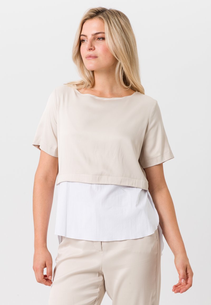 Beige top met korte mouwen en een gelaagd ontwerp, met een gladde textuur aan de bovenkant en een witte, licht gekreukte stof eronder.