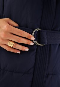 Abrigo acolchado azul marino con un cinturón texturizado que presenta un anillo metálico plateado. Una mano con un anillo dorado en capas descansa sobre el cinturón.