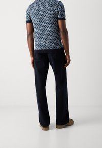 Mężczyzna w granatowej koszuli z krótkim rękawem w wzory, ciemnych spodniach szerokich nogawkach i beżowych butach na grubym spodzie, stojący na tle jasnego, jednolitego tła.