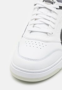 Białe skórzane trampki o gładkiej fakturze, z perforacjami na czubku, białymi sznurówkami i czarnym logo Pumy po boku.