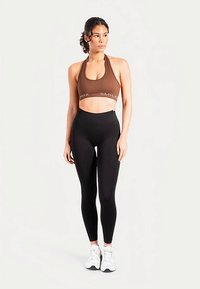 Brązowy sportowy stanik na ramiączkach z logo "SMIL", w połączeniu z czarnymi legginsami z wysokim stanem. Modelka ma na sobie białe buty sportowe, stoi na tle o neutralnym kolorze.