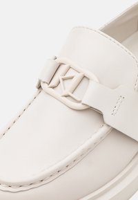 Mocassins en cuir blanc avec une boucle proéminente portant un logo, texture lisse et couture contrastante le long des bords.