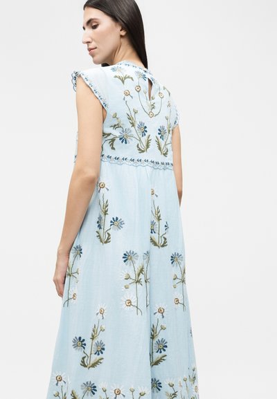 Γυναίκα με ανοιχτό γαλάζιο φόρεμα κεντημένο με λευκά και μπλε λουλούδια, με ραμμένα κοντά φουσκωτά μανίκια και άνοιγμα στο λαιμό στην πλάτη.