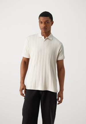 Texturiertes cremefarbenes Poloshirt mit einem gerippten Design, kurzen Ärmeln und einem traditionellen Kragen, kombiniert mit schwarzen Hosen.
