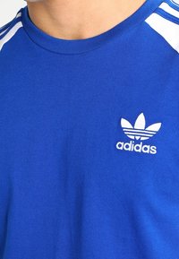 Sininen puuvillapaita, jossa on pyöreä kaulus. Olkapäillä valkoiset kolmonauha-akcentit ja rinnassa pieni valkoinen Adidas-logo.