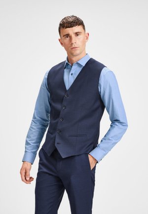 Gilet marine au tissu lisse, doté de cinq boutons, porté sur une chemise bleu clair à col. Coupe ajustée avec un léger éclat.